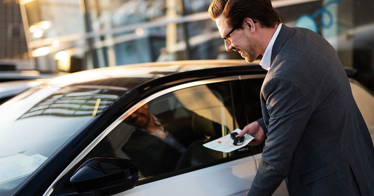 Ein Mann steht an einem Auto und hat den Schlüssel und Fahrzeugschein in der Hand. Er will gerade einsteigen und greift mit der anderen Hand zum Türgriff.