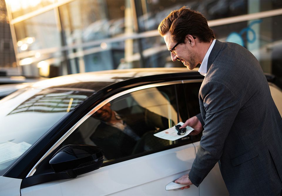 Ein Mann steht an einem Auto und hat den Schlüssel und Fahrzeugschein in der Hand. Er will gerade einsteigen und greift mit der anderen Hand zum Türgriff.
